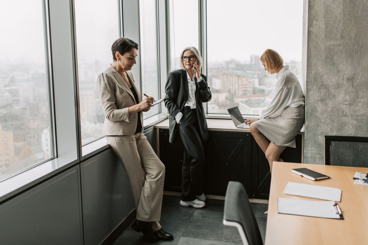Three businesswomen in corporate attire having a meeting in a modern office with a city view.