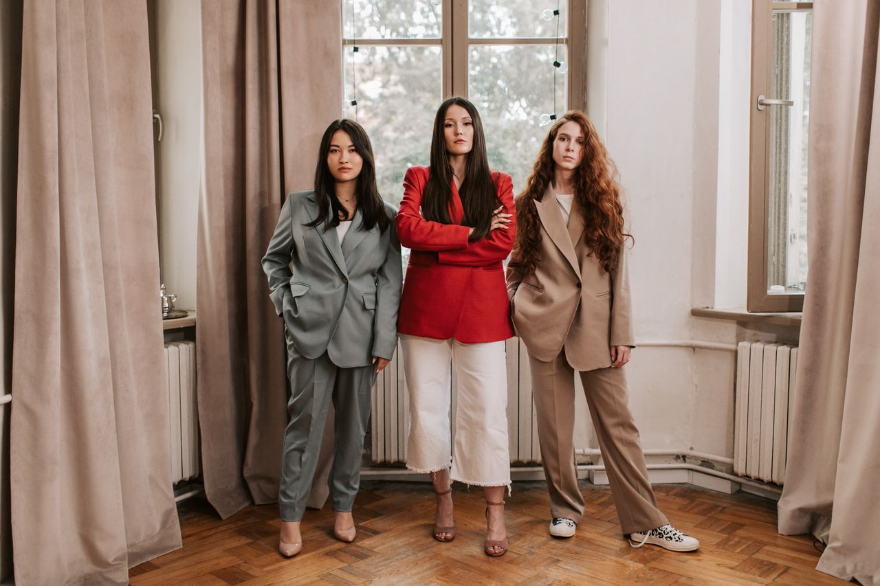 Home Three women confidently pose indoors, showcasing stylish business attire against elegant curtains.